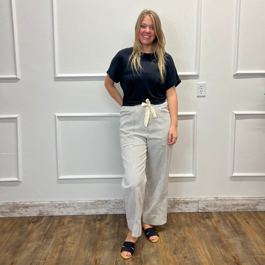 A woman stands in a room with white paneled walls and a wood floor, wearing a black short-sleeve t-shirt, light beige wide-leg pants with a cream tie, and black slide sandals.