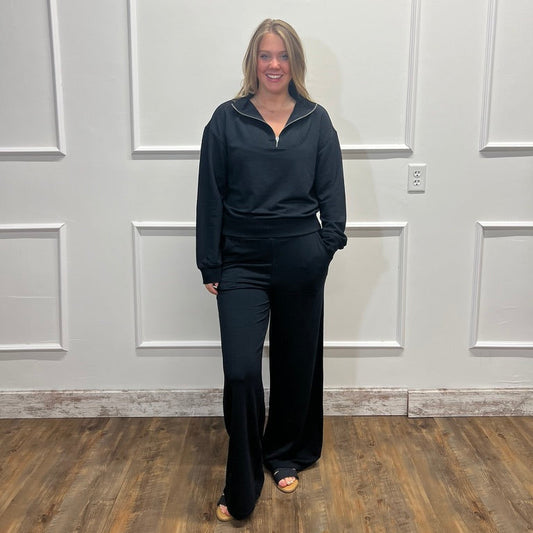 A woman stands indoors wearing a matching dark navy lounge set consisting of a half-zip long-sleeve top and wide-leg pants, set against a white paneled wall and wood-look floor.