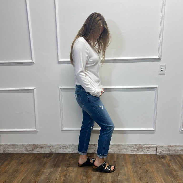 A woman stands in profile wearing a white long-sleeve top, blue skinny jeans, and black slide sandals, standing against a white paneled wall on a wood-look floor.