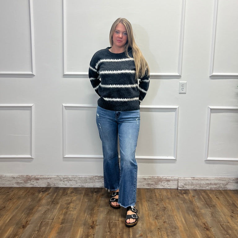 A woman stands in a room with white paneled walls and a wood floor, wearing a black and white patterned knit sweater, blue jeans, and black sandals.