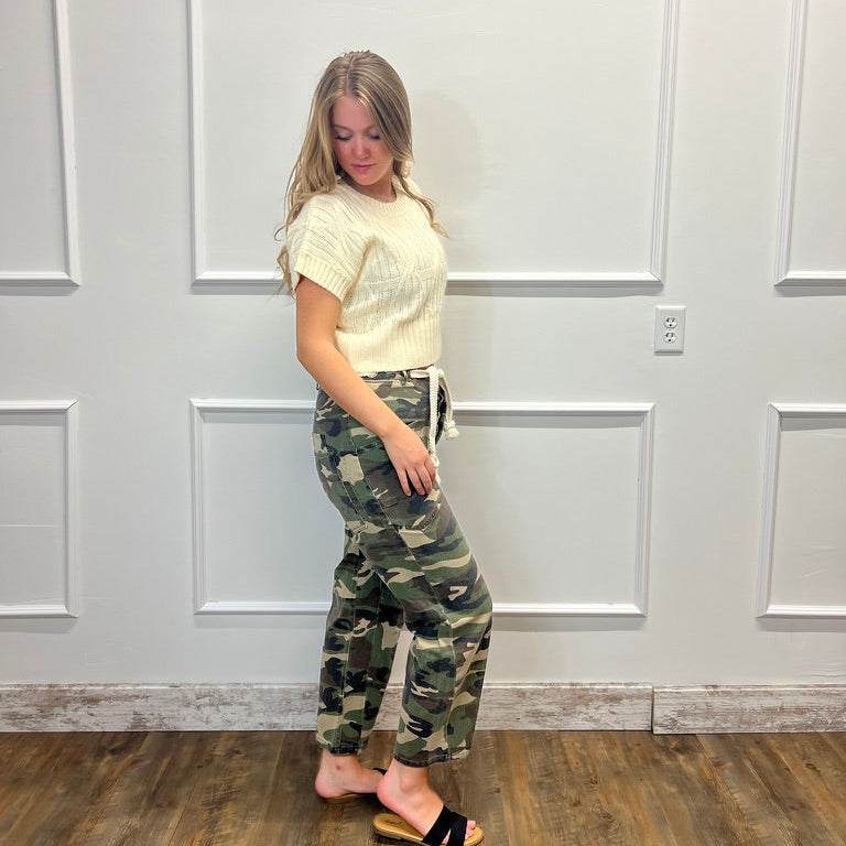 A woman stands in a room with white paneled walls and a wood floor, wearing a cream short-sleeve knit top and camouflage drawstring pants with black slide sandals.