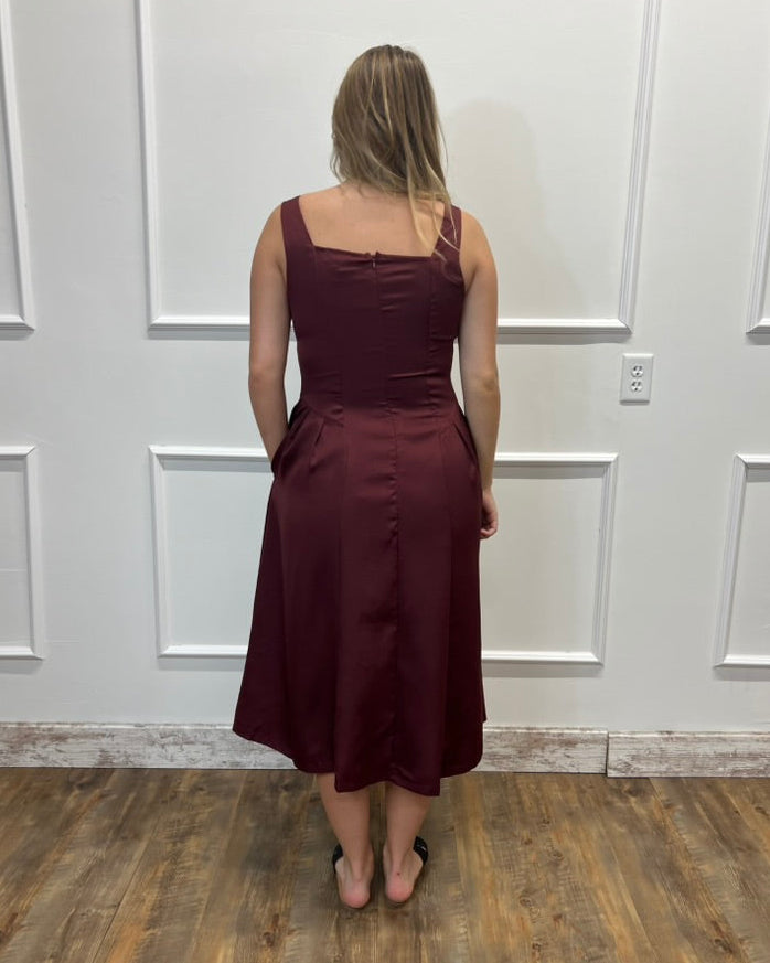 A woman viewed from behind wearing a sleeveless, knee-length burgundy dress with a fitted bodice and flared skirt, standing on a wood floor against a white paneled wall.