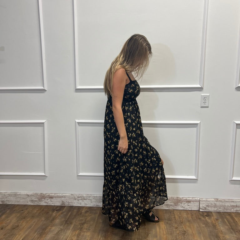A woman wearing a black maxi dress with a small yellow and white floral pattern, standing in a room with white paneled walls and a wood floor.