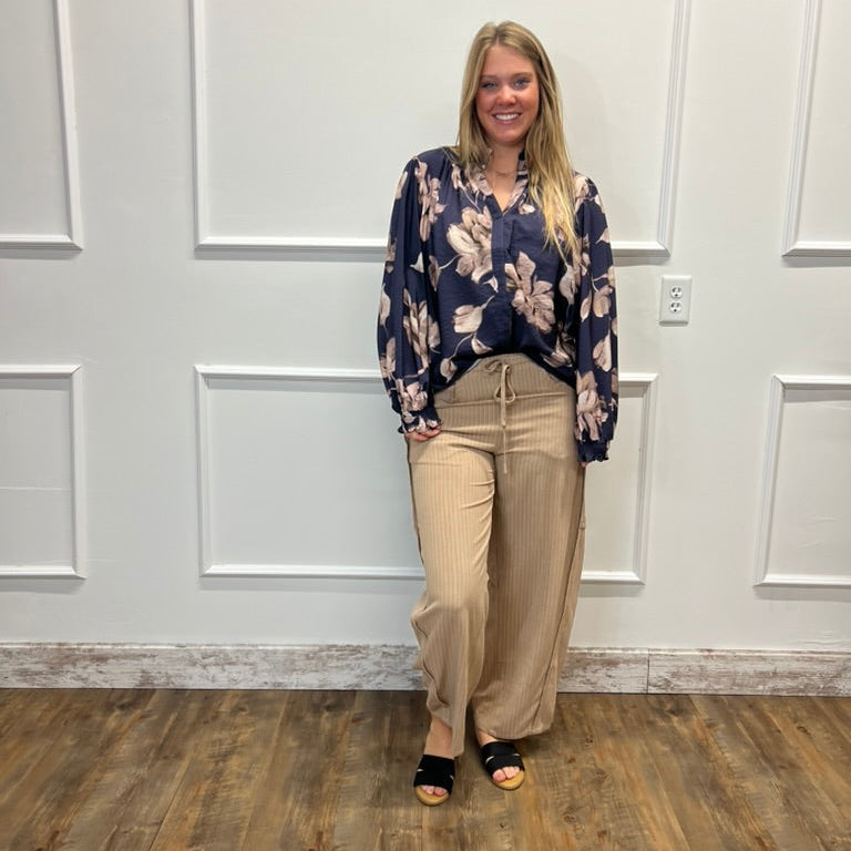 A woman stands in a room with white paneled walls and a wood floor, wearing a navy blue floral long-sleeve blouse and beige wide-leg pants with black sandals.