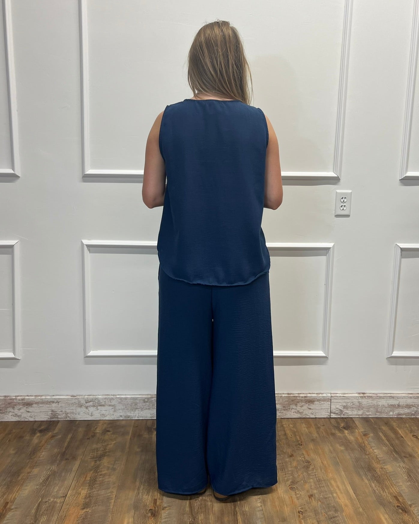 A woman viewed from behind wearing a sleeveless navy blue top and matching wide-leg pants, standing in front of a white paneled wall on a wood floor.