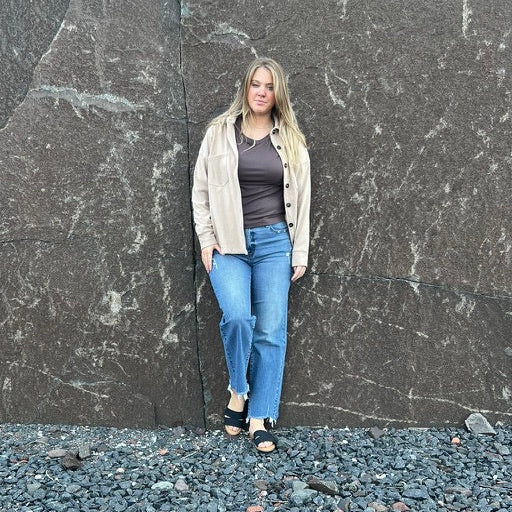 A woman stands in front of a massive dark stone wall on a bed of dark gravel, wearing a beige zip-up cardigan over a dark top and blue jeans.