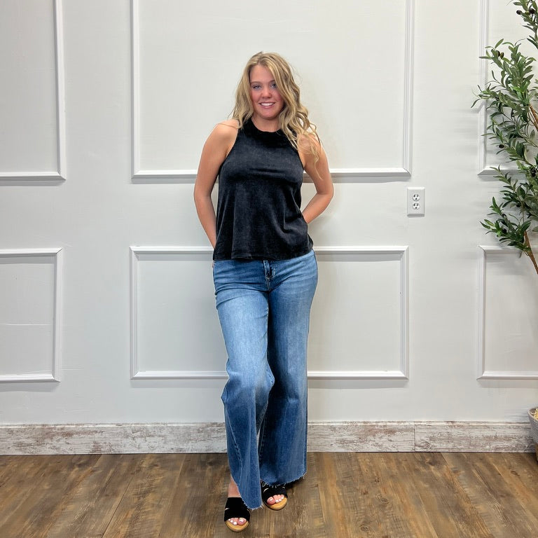 Woman wearing a black sleeveless top and blue jeans standing in a room with white paneled walls and wooden flooring.