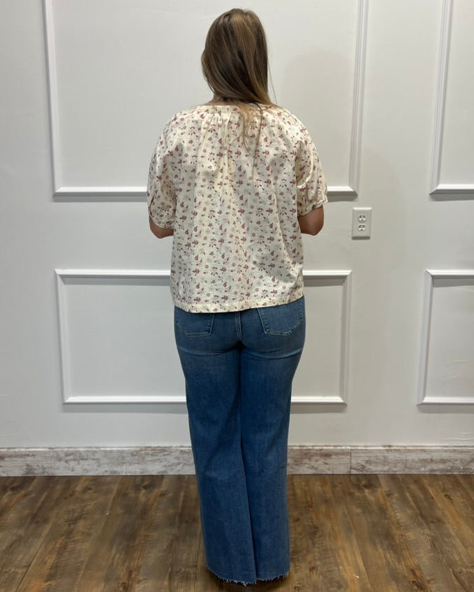 A woman viewed from behind wearing a cream-colored short-sleeve blouse with a small floral pattern and blue wide-leg jeans, standing in front of a white paneled wall on a wood floor.