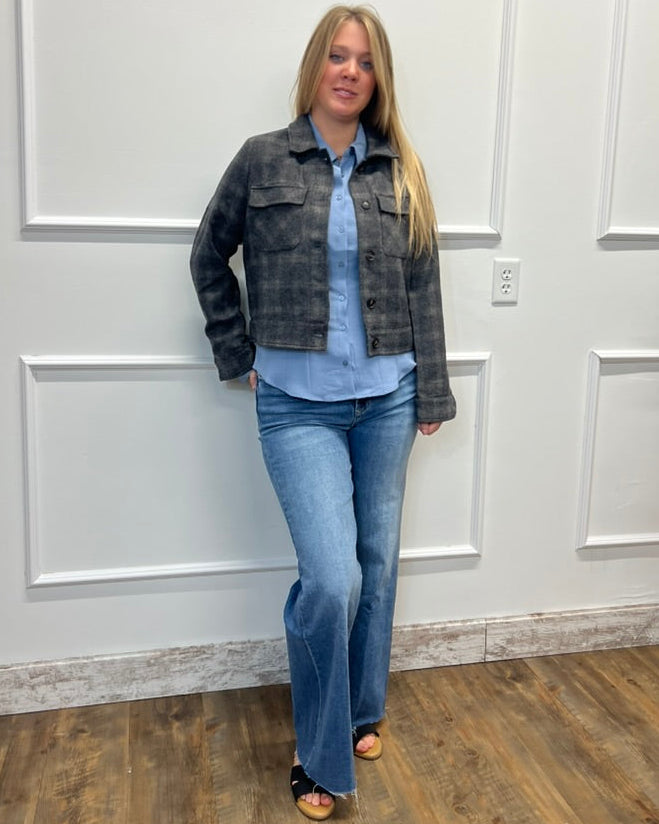 A woman modeling a dark gray plaid denim jacket over a light blue button-down shirt and blue jeans, standing against a white paneled wall on a wood floor.