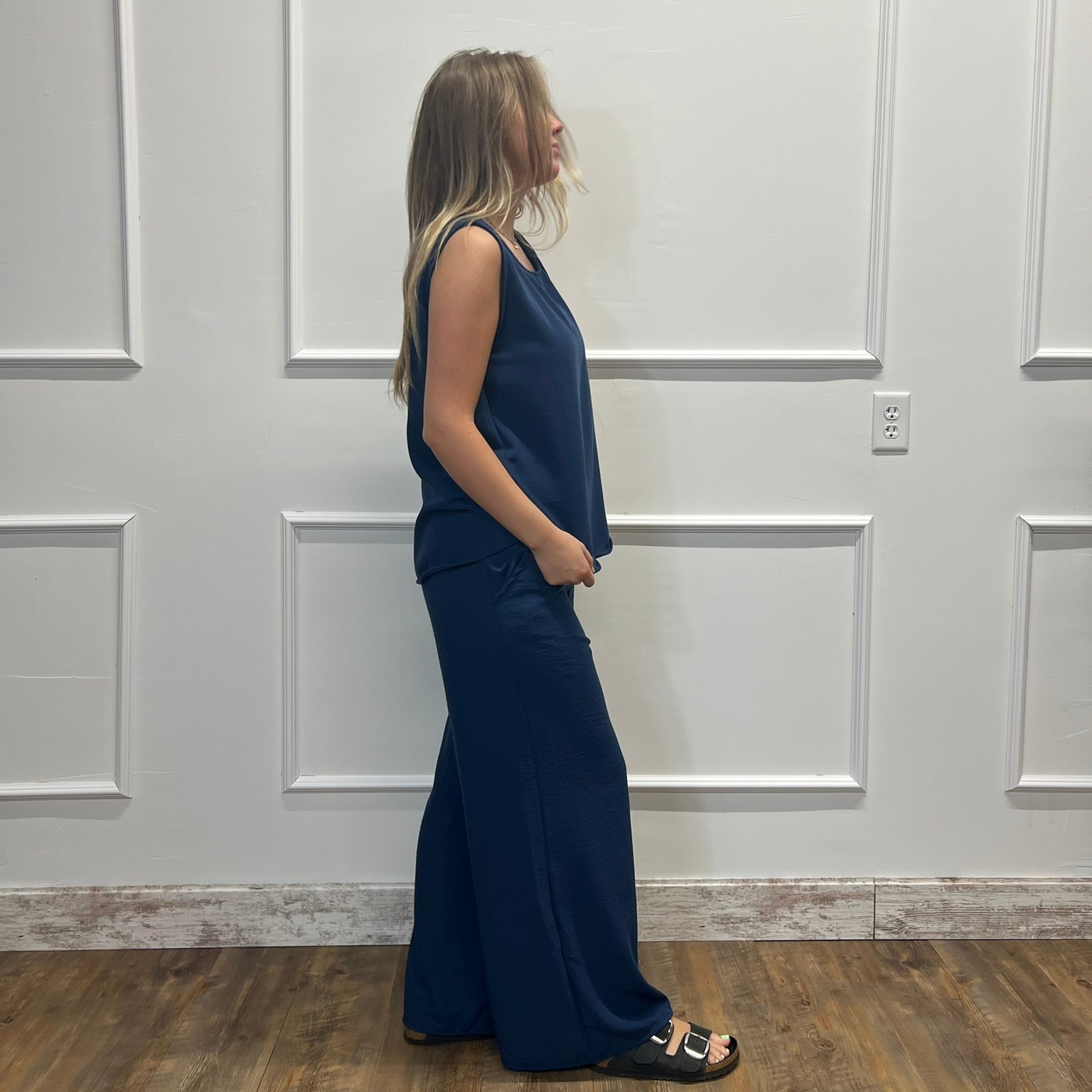 A woman stands in profile wearing a sleeveless navy blue maxi dress with a V-neckline and wide-leg pants, paired with black slide sandals, set against a white paneled wall and wood floor.