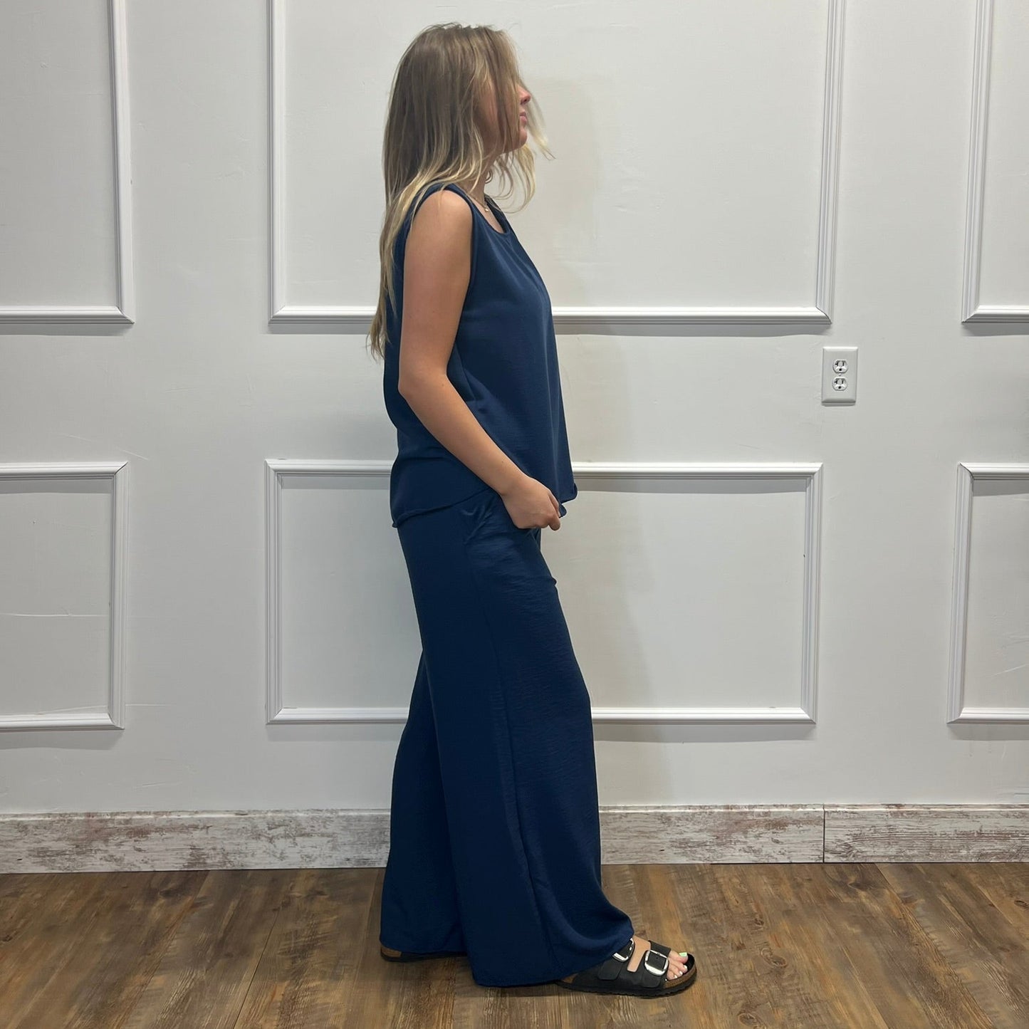 A woman stands in profile wearing a sleeveless navy blue maxi dress with a V-neckline and wide-leg pants, paired with black slide sandals, set against a white paneled wall and wood floor.