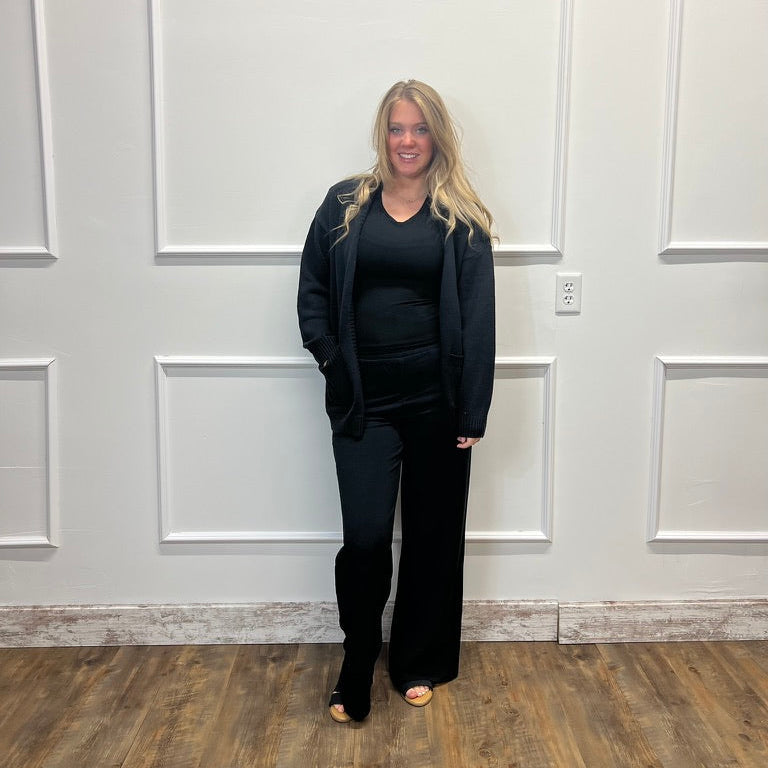 A woman with blonde hair stands in front of a white paneled wall wearing a black outfit consisting of a black top, black cardigan, and black wide-leg pants, with black open-toe heels.