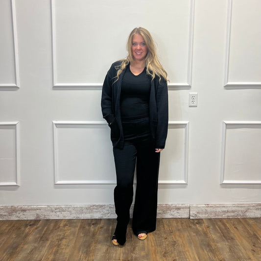 A woman with blonde hair stands in front of a white paneled wall wearing a black outfit consisting of a black top, black cardigan, and black wide-leg pants, with black open-toe heels.