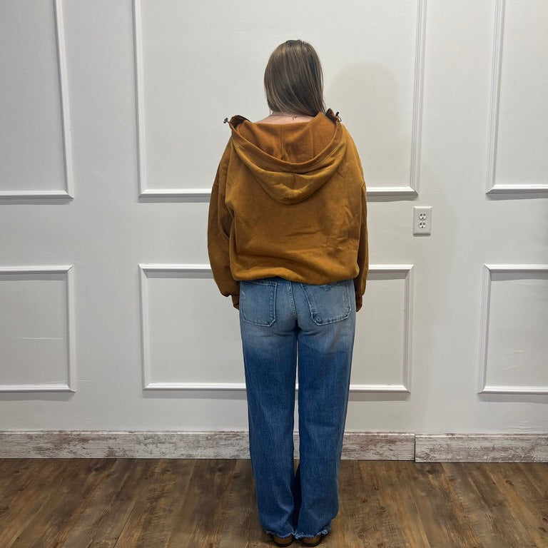 A person viewed from behind wearing a mustard yellow hooded sweatshirt and blue jeans, standing on a wood floor against a white paneled wall.
