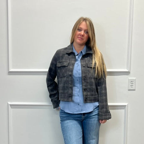 A woman stands indoors against a white paneled wall on a wood floor, wearing a dark washed denim jacket over a light blue button-up shirt and blue jeans.