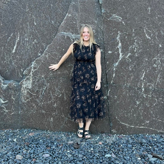 A woman stands in front of a massive dark stone wall outdoors, wearing a sleeveless black midi dress with a small floral pattern and black sandals.