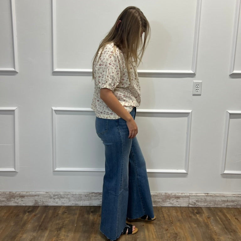 A woman stands in profile wearing a cream short-sleeve blouse with a small floral print and blue flare jeans, standing against a white paneled wall on a wood floor.