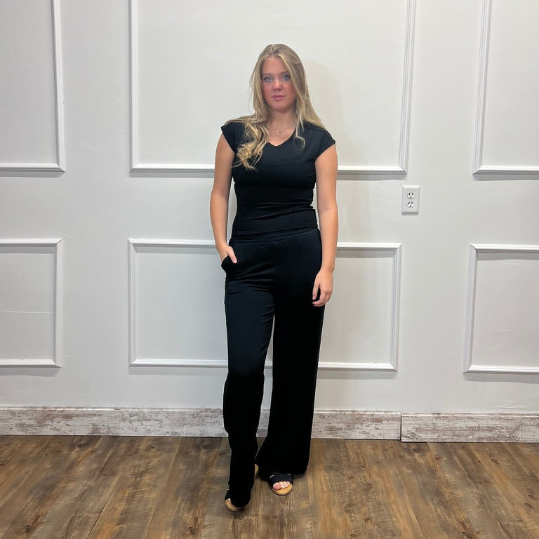A woman modeling a black sleeveless jumpsuit with a V-neckline and wide-leg pants, standing in front of a white paneled wall on a wood floor.