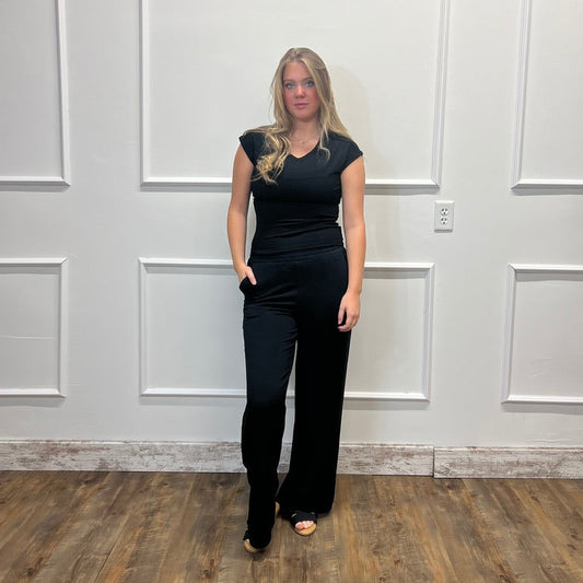 A woman modeling a black sleeveless jumpsuit with a V-neckline and wide-leg pants, standing in front of a white paneled wall on a wood floor.