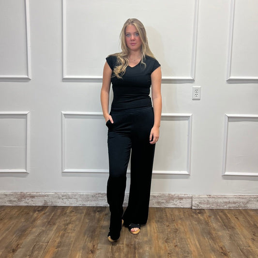 A woman modeling a black sleeveless jumpsuit with a V-neckline and wide-leg pants, standing in front of a white paneled wall on a wood floor.