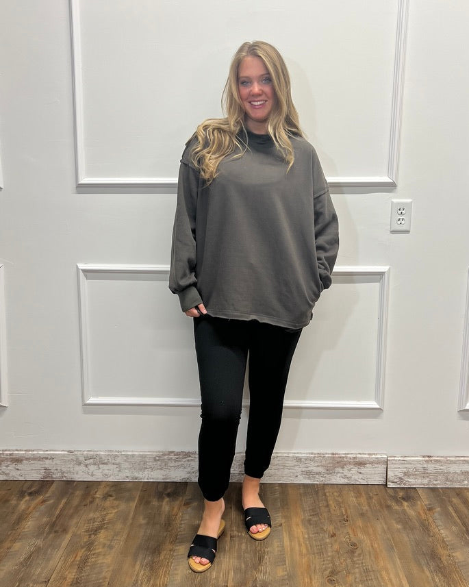 A woman stands indoors against a white paneled wall wearing an oversized dark gray long-sleeve top, black pants, and black slide sandals.