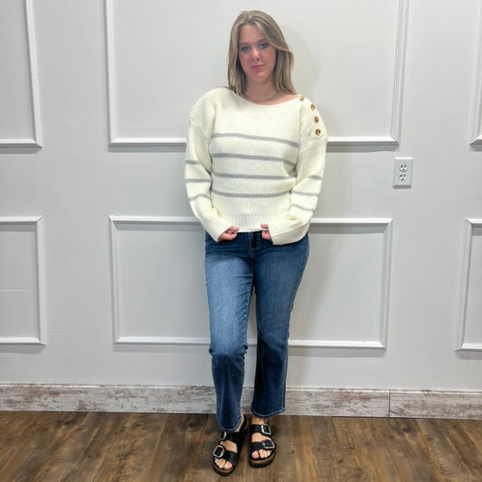 A woman stands in front of a white paneled wall wearing a cream knit sweater with horizontal gray stripes and gold button details on the sleeves, paired with blue jeans and black sandals.