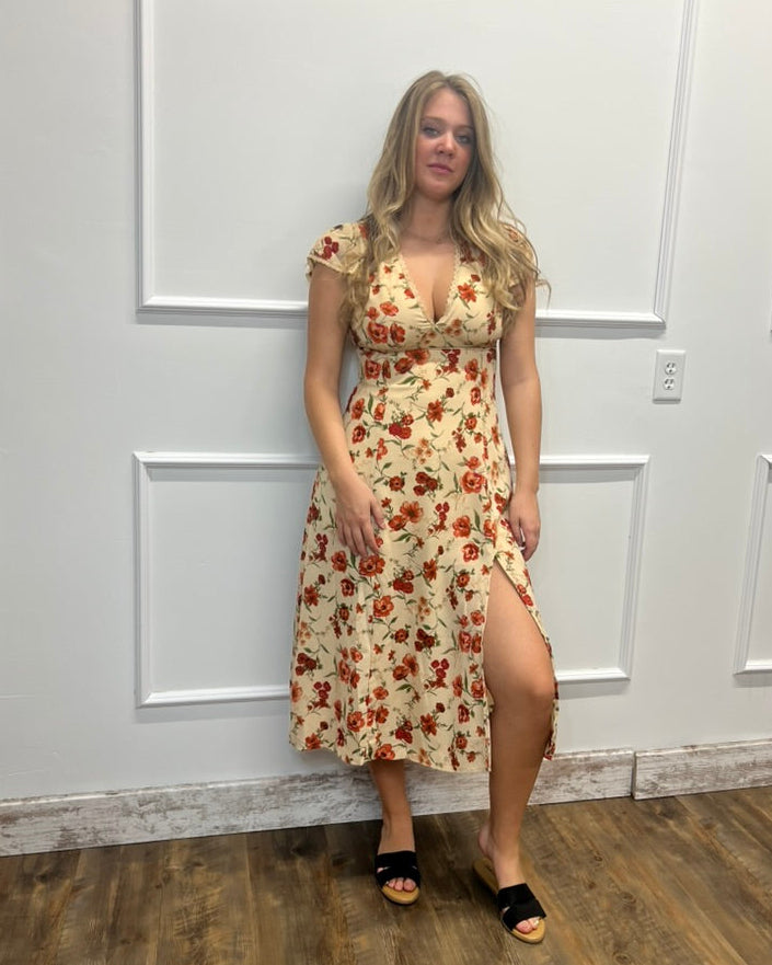 A woman modeling a cream-colored midi dress with a red and orange floral pattern, standing against a white paneled wall on a wood floor.