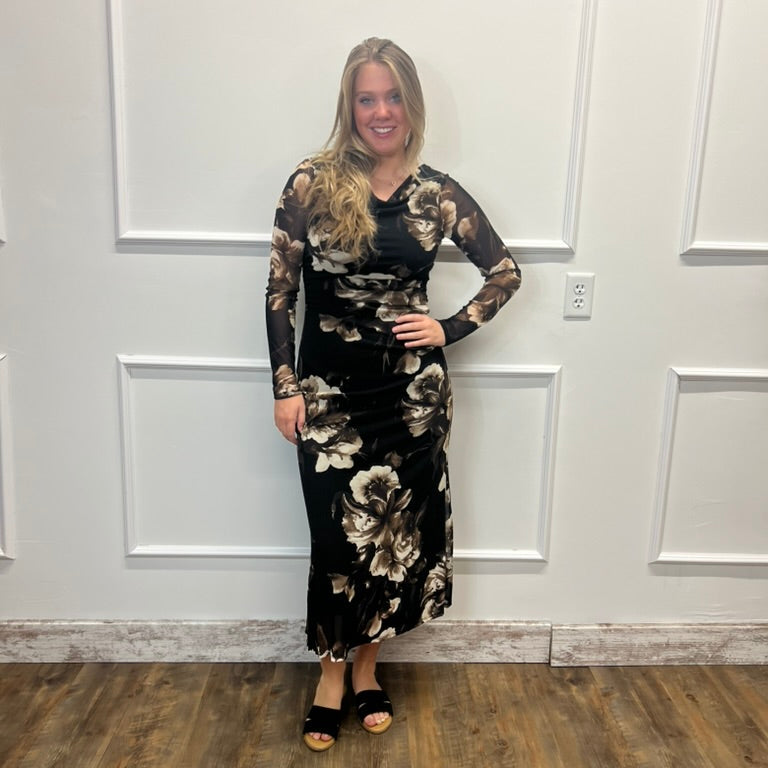 A woman modeling a black long-sleeve maxi dress with a beige and white floral pattern, standing against a white paneled wall on a wood floor.