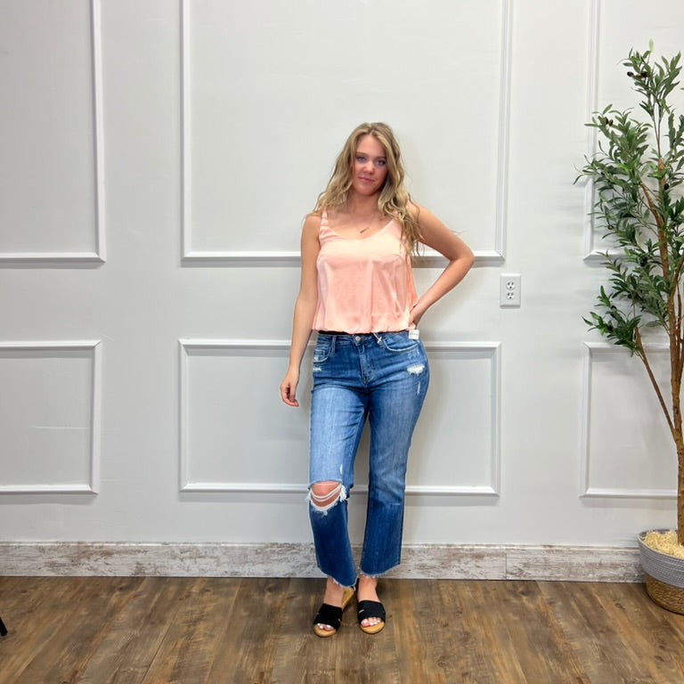 Woman wearing a pink top and blue jeans standing in a room with a white paneled wall and a plant.
