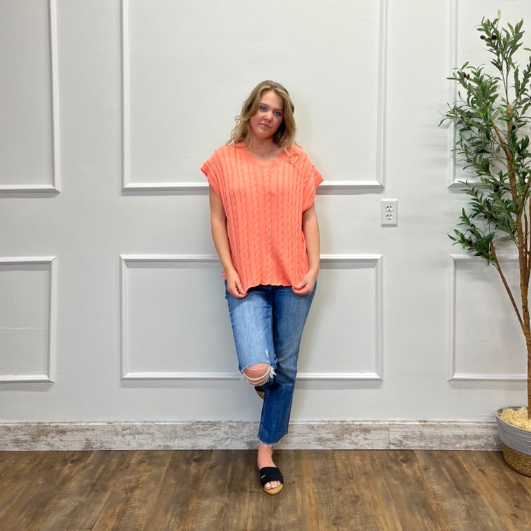 Woman in an orange top and blue jeans standing against a white paneled wall.