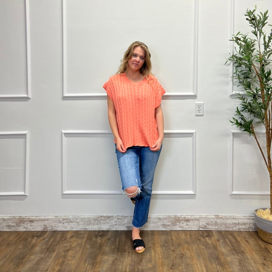 Woman in an orange top and blue jeans standing against a white paneled wall.