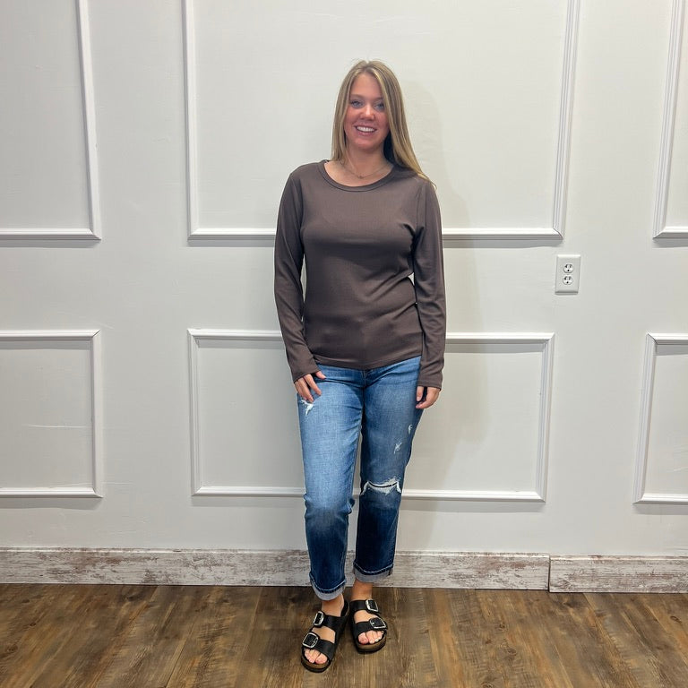 A woman stands in a room with white paneled walls and a wood floor, wearing a dark brown long-sleeve crewneck top, blue ripped jeans, and black sandals.