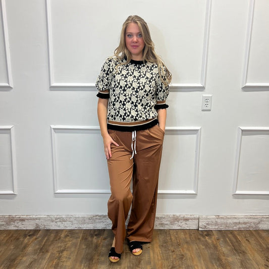 A woman stands in a room with white paneled walls and a wood floor, wearing a black and white patterned short-sleeve top, brown wide-leg pants, and black sandals.