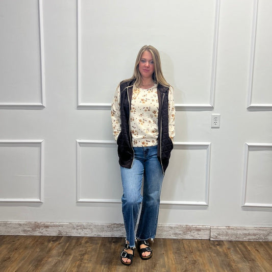 A woman stands against a white paneled wall wearing a cream floral long-sleeve top, a black zippered vest, blue jeans, and black sandals.