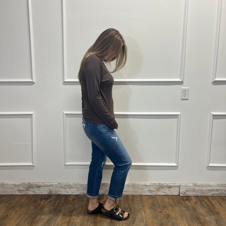 A woman stands in profile wearing a dark brown long-sleeve top, blue jeans, and black slide sandals with gold hardware, set against a white paneled wall and wood-look flooring.