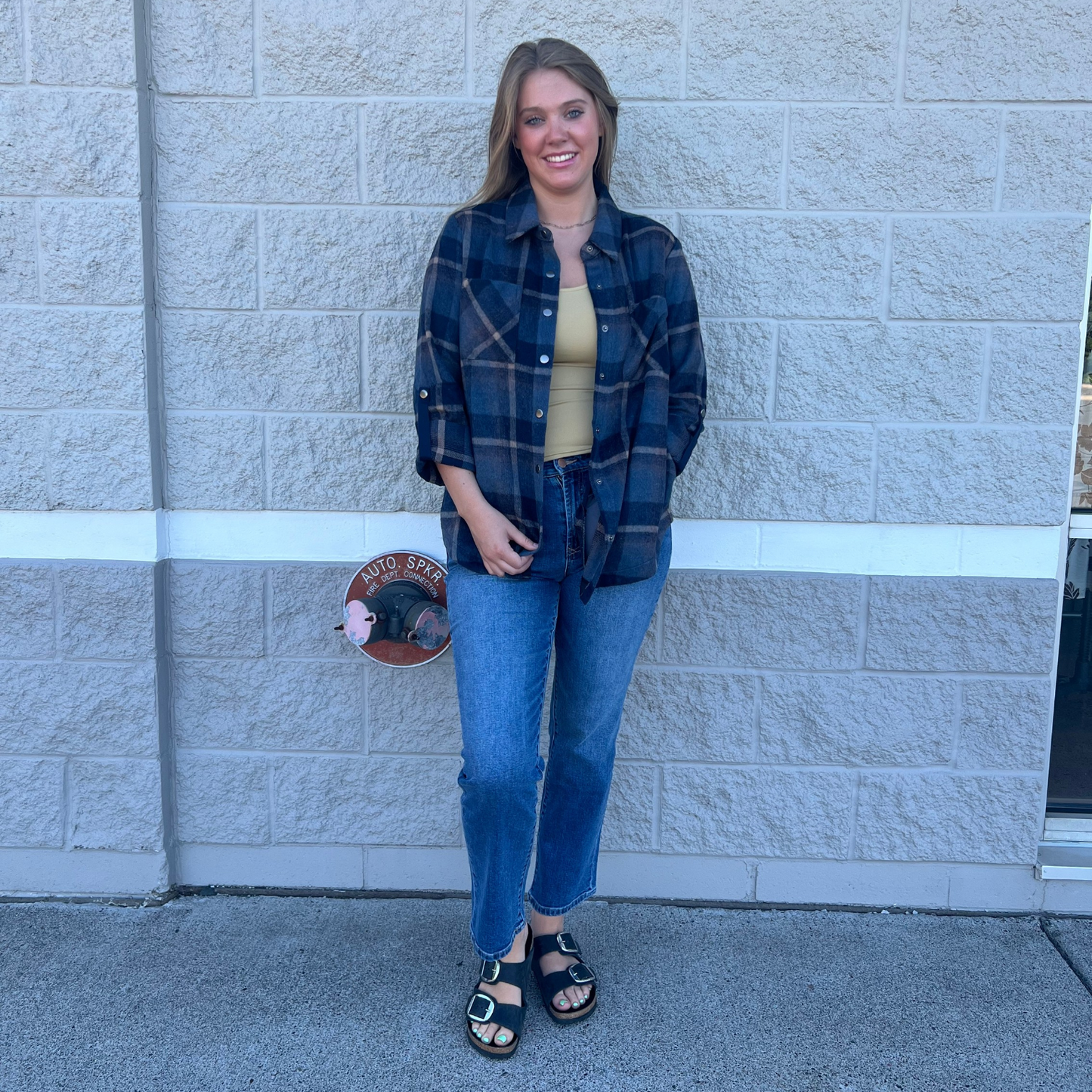 A woman stands against a light gray stone wall wearing a dark blue and black plaid button-up shirt over a beige top, blue jeans, and black sandals.