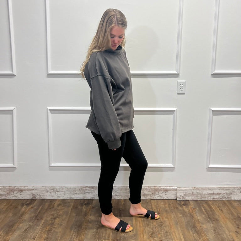 A woman stands in profile wearing a loose gray sweatshirt, black leggings, and black slide sandals, standing on a wood-look floor against a white paneled wall.