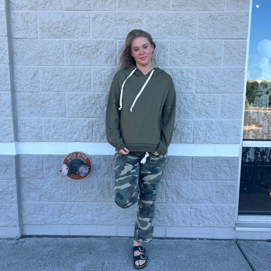 A woman stands against a light gray brick wall wearing an olive green hoodie with white drawstrings and camouflage pants, paired with black slide sandals.