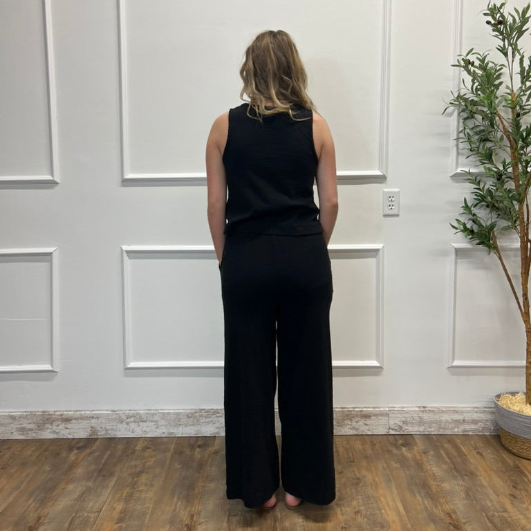 Person wearing a black sleeveless top and wide-leg pants standing in a room with white paneled walls and a plant.