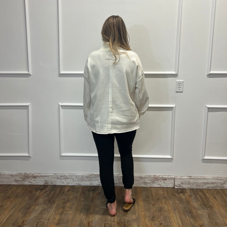 A woman viewed from behind wearing a loose white long-sleeve button-up shirt, black pants, and black sandals, standing in front of a white paneled wall on a wood floor.