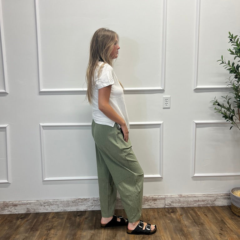 A woman stands in profile wearing a white short-sleeve t-shirt and olive green wide-leg pants, paired with black slide sandals, in front of a white paneled wall with a wooden floor.