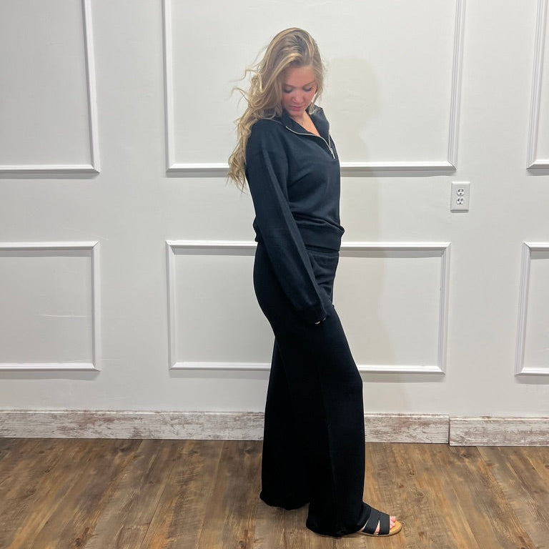 A woman stands in profile wearing a black long-sleeve collared top and matching wide-leg pants against a white paneled wall with wood flooring.