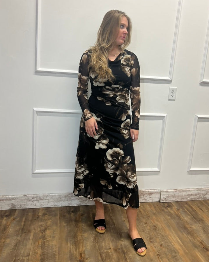 A woman modeling a long-sleeve midi dress with a black base and a brown and beige floral pattern, standing in front of a white paneled wall on a wood floor.