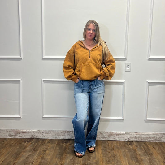 A woman stands against a white paneled wall wearing a mustard yellow zip-up hoodie and light blue wide-leg jeans.