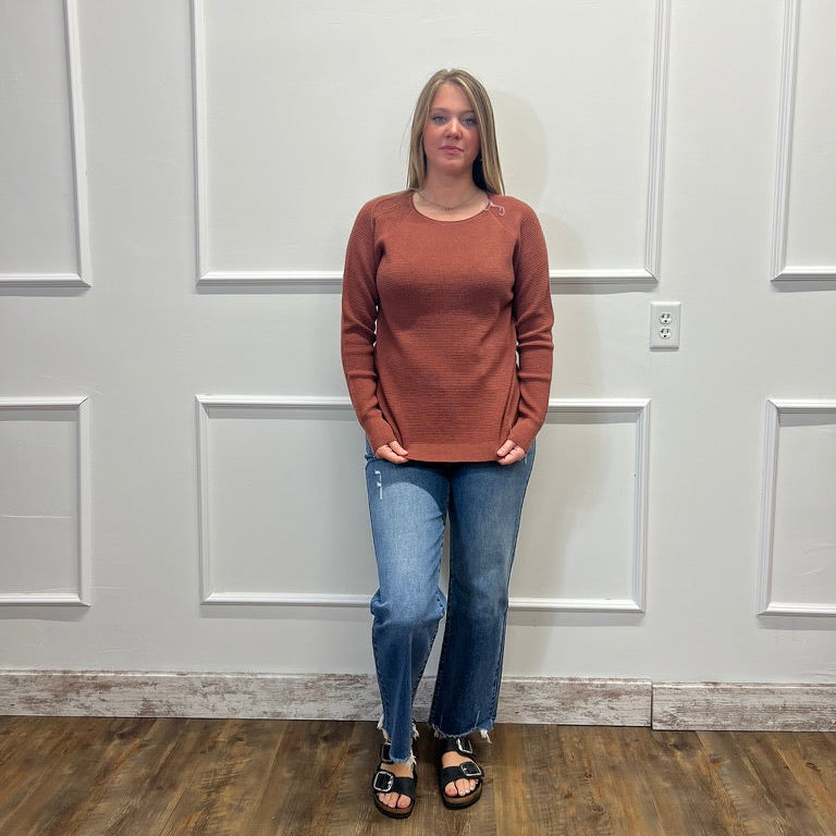 A woman stands in a room with white paneled walls and wood floors, wearing a rust-brown long-sleeve crewneck sweater, blue jeans, and black sandals.