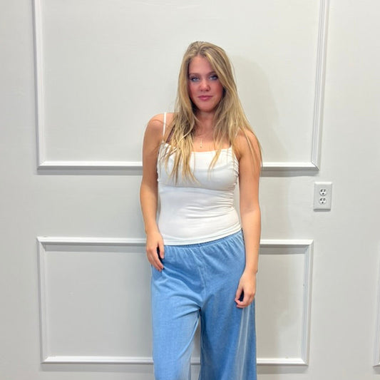 A woman stands indoors against a white paneled wall wearing a white sleeveless top and light blue wide-leg pants, with white slide sandals.