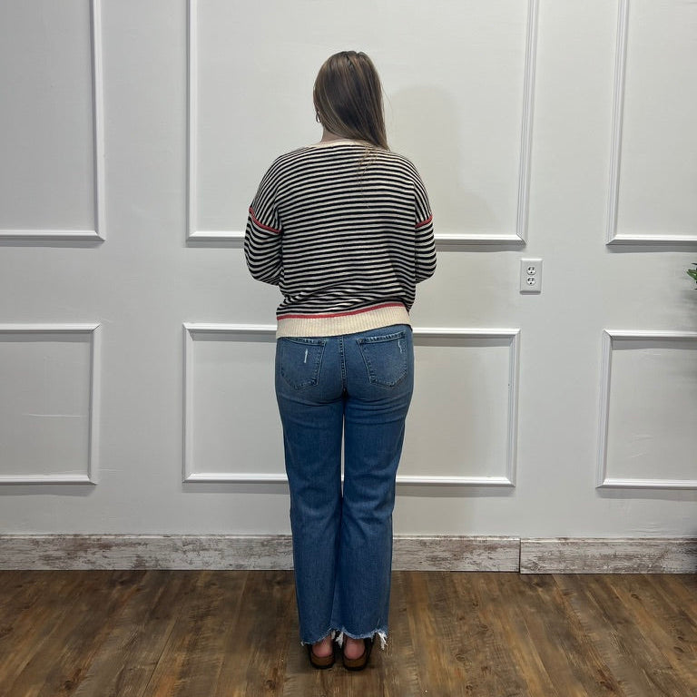 A woman viewed from behind wearing a black and white striped long-sleeve crop top and blue jeans, standing in front of a white paneled wall on a wood floor.