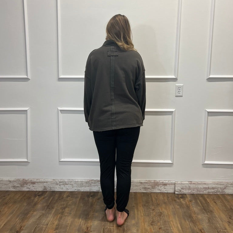 A woman viewed from behind wearing a dark olive green button-up shirt, black pants, and pink slip-on shoes, standing on a wood floor against a white paneled wall.