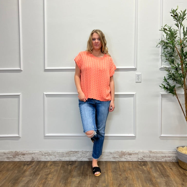 Woman in an orange top and blue jeans standing against a white paneled wall.