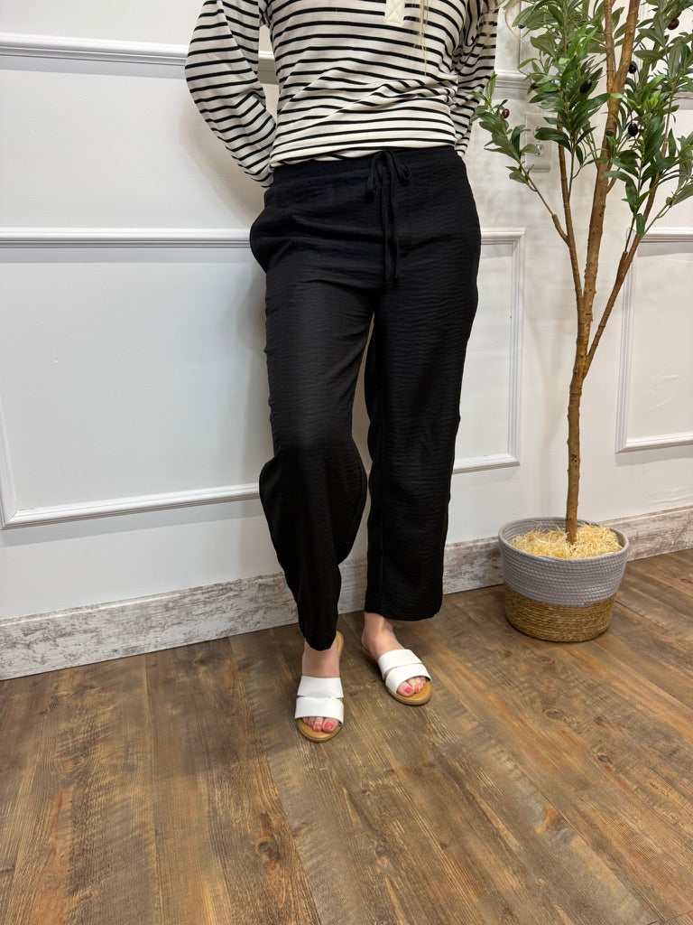 Person wearing a striped shirt and black pants standing against a white paneled wall with a plant in the background.